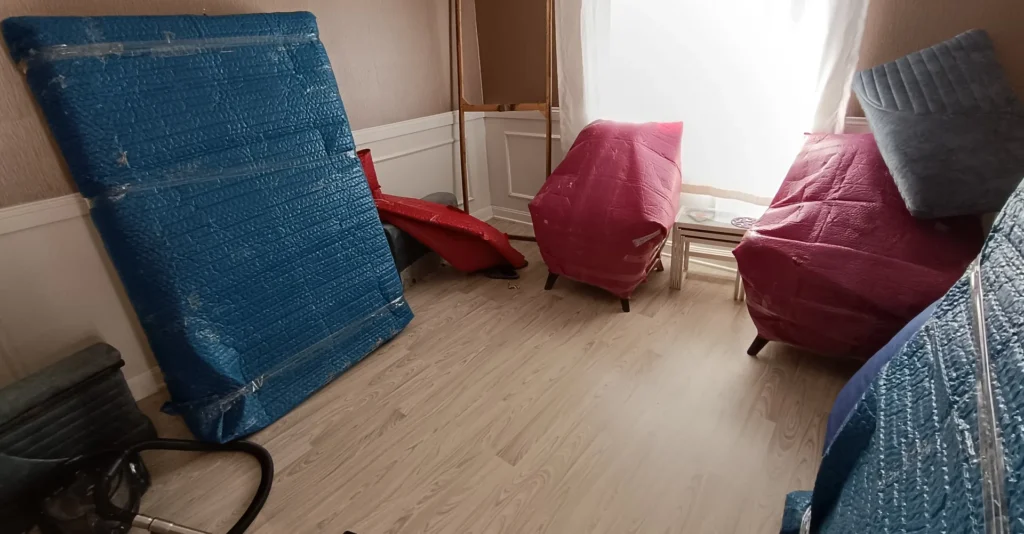 Mattress and armchairs wrapped in colored moving blankets and tape in a bright room