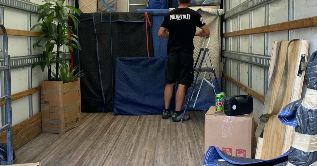 A mover with his back to the camera loading blanket-wrapped furniture inside a box truck
